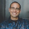 A man with glasses and short dark hair smiles while standing against a dark wooden door.