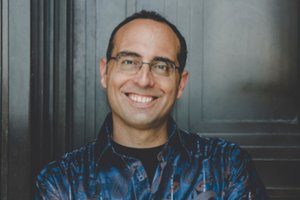 A man with glasses and short dark hair smiles while standing against a dark wooden door.