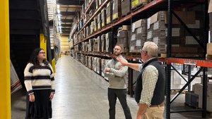 Three people stand and talk in a large warehouse aisle lined with tall shelves stacked with cardboard boxes.