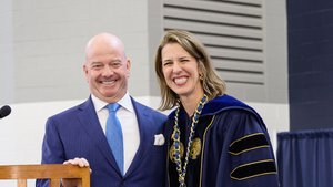 A man in a blue suit and a woman in dark blue academic regalia with a gold presidential chain smile during a university