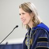 A woman in a blue academic gown with a gold chain smiles warmly while speaking into a microphone.