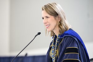 A woman in a blue academic gown with a gold chain smiles warmly while speaking into a microphone.