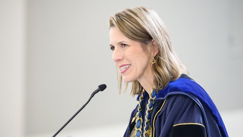 A woman in a blue academic gown with a gold chain smiles warmly while speaking into a microphone.