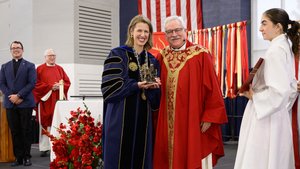 A woman in academic gown holds an ornate object with a smiling priest in red vestments beside her at a university ceremony.