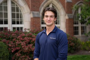 A young man wearing a navy blue John Carroll University pullover stands smiling in front of a brick building with arched.