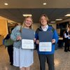 A smiling young woman and man stand indoors holding open certificate folders showing their awards.