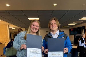 A smiling young woman and man stand indoors holding open certificate folders showing their awards.