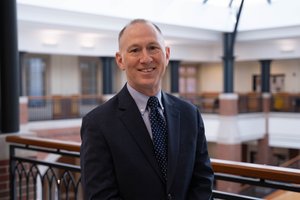 A warm smile lights the face of a man in a navy suit and light blue shirt, standing in a bright building atrium.