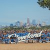 A large group of people wearing blue shirts pose outdoors behind a white Cleveland sign with a city skyline in the