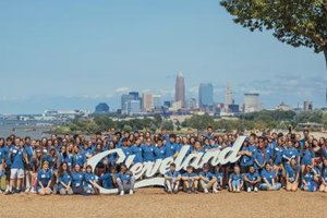 A large group of people wearing blue shirts pose outdoors behind a white Cleveland sign with a city skyline in the