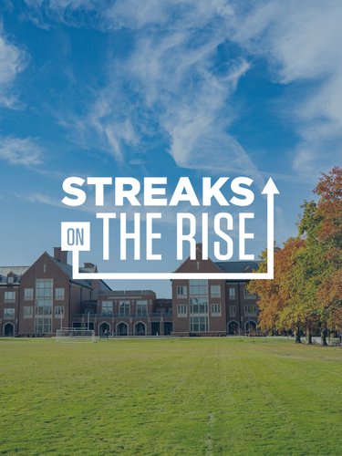 A large brick building sits behind a wide grassy field under a blue sky with wispy clouds and autumn trees on the right.