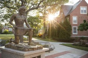 A bronze statue of a kneeling man holding a long object is in front of a brick building with sunlight shining through trees.
