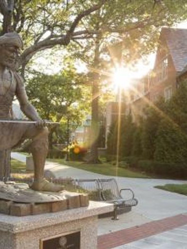 A bronze statue of a kneeling man holding a long object is in front of a brick building with sunlight shining through trees.
