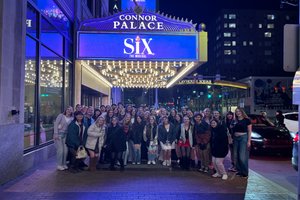 A large group of people pose smiling under the illuminated Connor Palace theater marquee for Six the Musical at night.