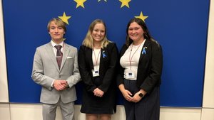 Three people dressed in business attire stand smiling in front of a blue wall with yellow stars arranged in a circle.
