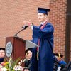A young man in a blue graduation gown and cap speaks at a podium decorated with flowers while seated faculty watch.