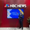 A man in a dark suit stands with arms crossed in front of a red wall displaying the NBC News logo and a screen showing Meet.