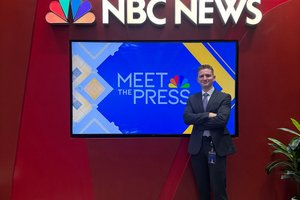 A man in a dark suit stands with arms crossed in front of a red wall displaying the NBC News logo and a screen showing Meet.