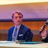 Two young men in suits sit at a table, one speaking into a microphone while the other listens attentively.