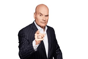 A bald man in a dark pinstripe suit points directly forward with a serious expression on his face.