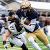 A football player in a navy jersey and gold pants runs with the ball while a player in a white jersey lunges to tackle him.