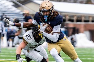 A football player in a navy jersey and gold pants runs with the ball while a player in a white jersey lunges to tackle him.