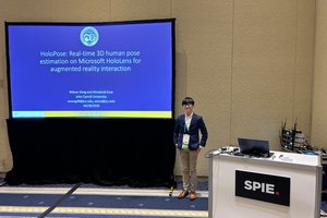 A man in a suit stands smiling next to a presentation screen titled HoloPose about 3D human pose estimation.