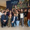 A group of twenty people pose indoors in front of a colorful John Carroll sign, with one person holding a framed certificate.
