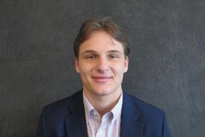 A young man smiles, wearing a navy blazer over a patterned shirt, against a dark gray background.