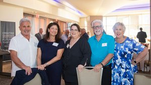 Five adults stand closely together smiling inside a well-lit room with large windows and a table with flowers in the