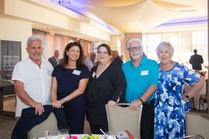 Five adults stand closely together smiling inside a well-lit room with large windows and a table with flowers in the