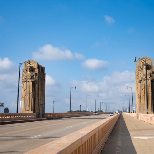 Cars drive over a bridge, with street lamps and two tall stone statues on either side under a blue sky.