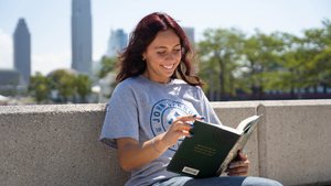 A student smiles, looking down at a book held open, while sitting outside.