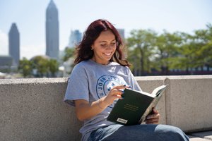 A student smiles, looking down at a book held open, while sitting outside.