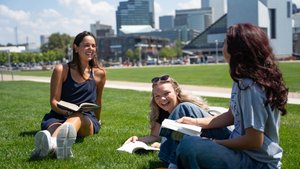 Three students sit on the grass and read their textbooks, smiling at each other; buildings stand in the background.