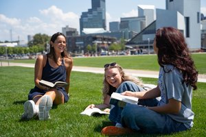 Three students sit on the grass and read their textbooks, smiling at each other; buildings stand in the background.