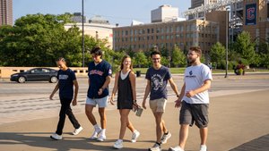 Five students walk together on a sidewalk; a ballpark and downtown buildings are visible in the background.