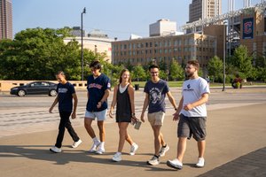 Five students walk together on a sidewalk; a ballpark and downtown buildings are visible in the background.