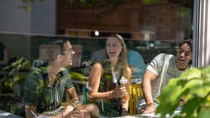 Three students at a cafe share laughs over ice cream, view through the window.