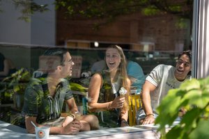 Three students at a cafe share laughs over ice cream, view through the window.