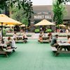 People relax at wooden picnic tables with yellow umbrellas, enjoying food from the Van Aken Market Hall.