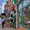 A person walks by Corbo's Bakery and Cafe, where flags hang outside and outdoor tables are set up.