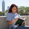 A student sits outside, smiling as they read a book on a sunny day in the city.
