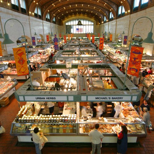 Shoppers browse food vendors at an indoor market, with the US flag visible in the background.