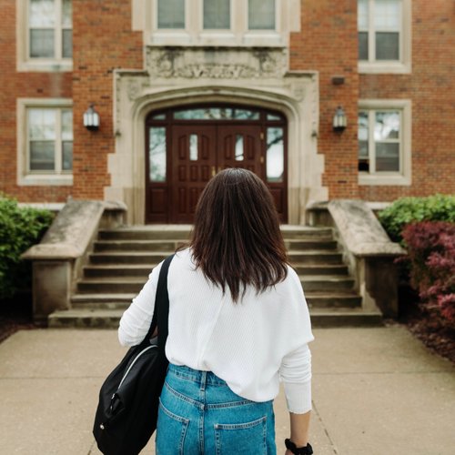 A person wearing a white sweater and denim skirt walks toward a brick building with stone steps and double wooden doors.