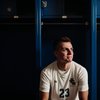 A young man in a white soccer jersey with number twenty-three sits in front of blue lockers looking to the side.