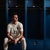 A male soccer player in a white uniform sits on a bench in front of blue lockers, looking to the side.