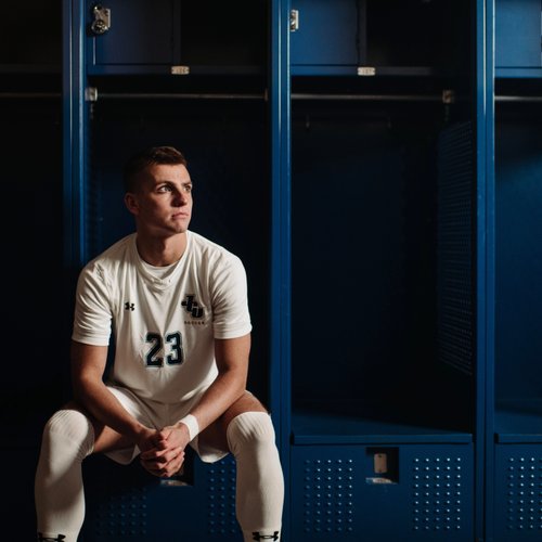 A male soccer player in a white uniform sits on a bench in front of blue lockers, looking to the side.