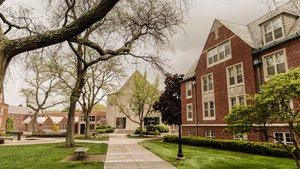 A paved walkway leads through a green lawn with trees toward a stone chapel and red brick buildings under a cloudy sky.