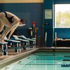 A swimmer in a black swimsuit and swim cap is poised on a starting block, ready to dive into an indoor pool.